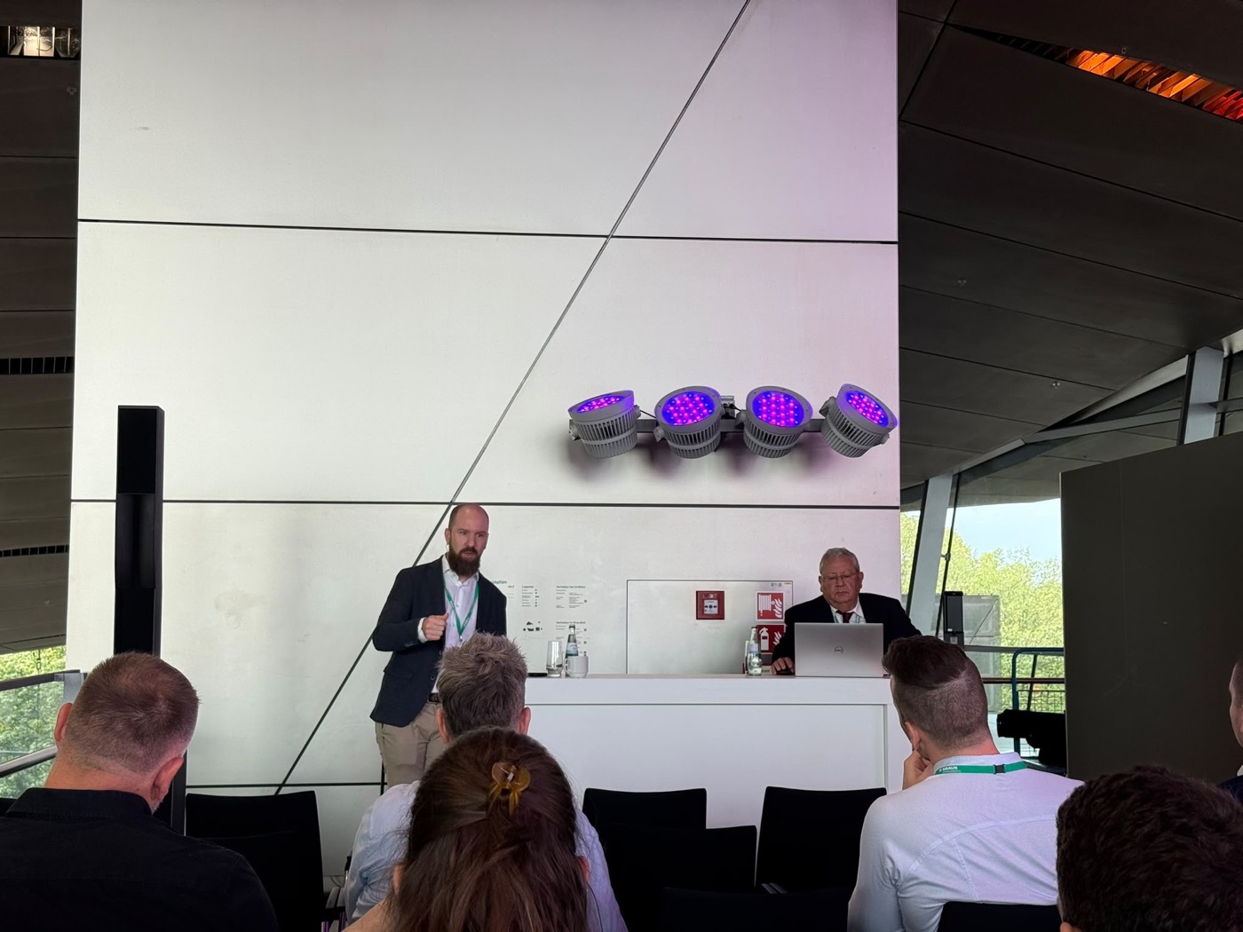 Matthias Verstraete standing to address a seated audience during the debate, with the session chair at the podium